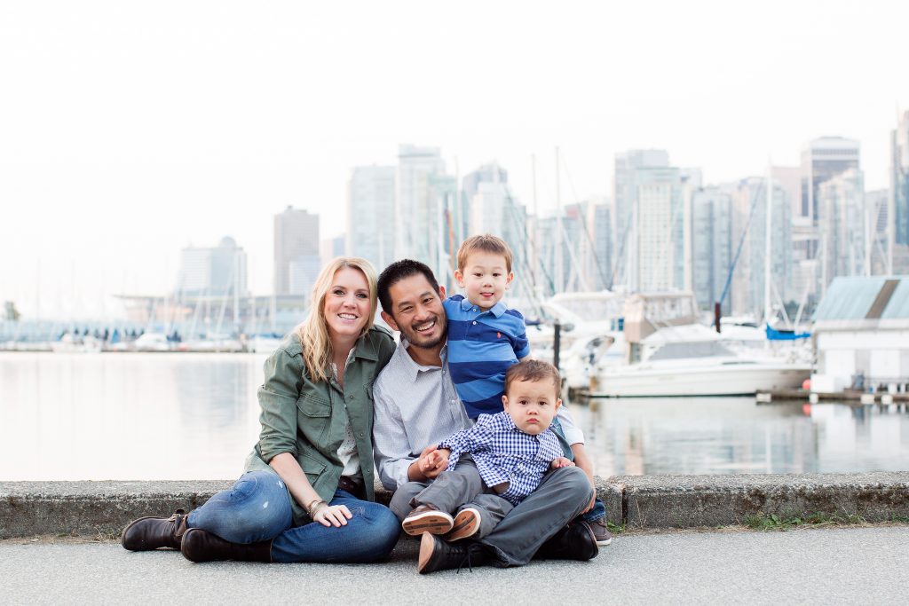 family of 4 sitting on the seawall in Stanley Park with the Vancouver skyline in the background